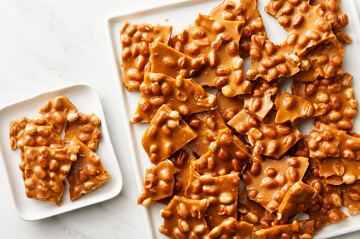 Pieces of golden-brown peanut brittle piled on a large white rectangular platter, with a smaller square plate containing a few more pieces to the left. Pieces of golden-brown peanut brittle piled on a large white rectangular platter, with a smaller square plate containing a few more pieces to the left.