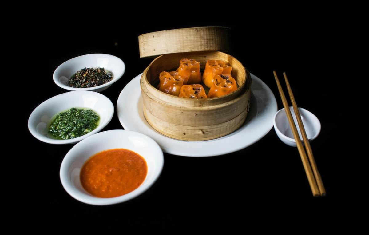 An open round bamboo steamer basket sits on a white plate, filled with five vibrant orange, open-top dim sum dumplings. Three small white bowls containing different colorful dipping sauces are arranged on the left, while a pair of wooden chopsticks rests on a tiny bowl to the right, all set against a solid black background. An open round bamboo steamer basket sits on a white plate, filled with five vibrant orange, open-top dim sum dumplings. Three small white bowls containing different colorful dipping sauces are arranged on the left, while a pair of wooden chopsticks rests on a tiny bowl to the right, all set against a solid black background.