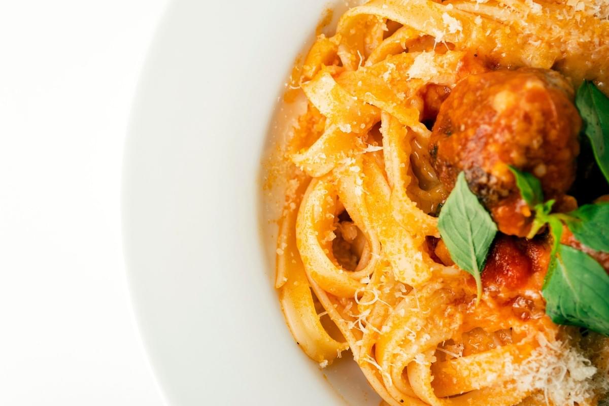 A top-down, close-up shot shows a white plate filled with a portion of fettuccine pasta tossed in a vibrant red tomato sauce. The dish is topped with a large meatball, sprinkled with grated parmesan cheese, and garnished with fresh green basil leaves. A top-down, close-up shot shows a white plate filled with a portion of fettuccine pasta tossed in a vibrant red tomato sauce. The dish is topped with a large meatball, sprinkled with grated parmesan cheese, and garnished with fresh green basil leaves.