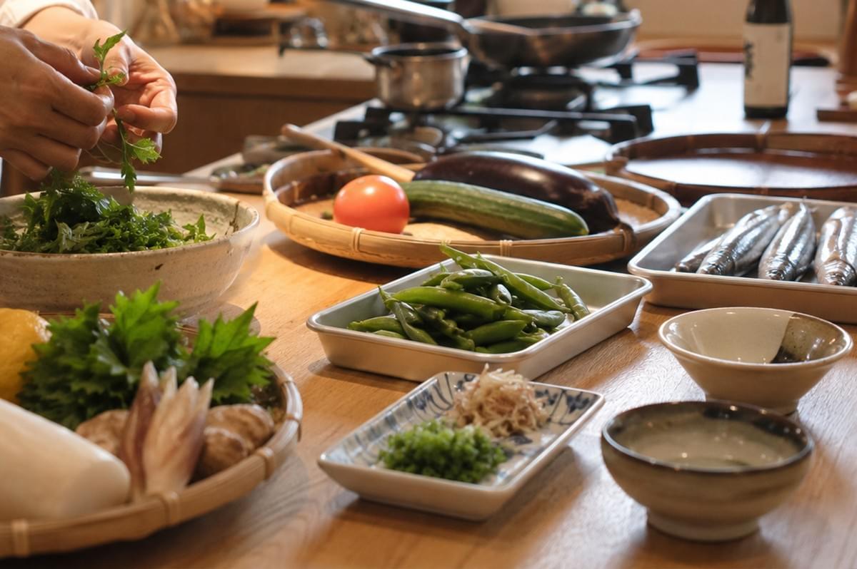 In a bright kitchen, a person’s hands are shown preparing ingredients, including a bowl of leafy greens being sorted and washed. The wooden counter is filled with a variety of fresh produce, including a large eggplant, a cucumber, a tomato, a metal tray of green beans, and a tray of whole fish waiting to be cleaned. Small bowls containing garnishes like shredded ginger and chopped scallions are arranged neatly, showcasing the meticulous preparation typical of Japanese culinary practices. In a bright kitchen, a person’s hands are shown preparing ingredients, including a bowl of leafy greens being sorted and washed. The wooden counter is filled with a variety of fresh produce, including a large eggplant, a cucumber, a tomato, a metal tray of green beans, and a tray of whole fish waiting to be cleaned. Small bowls containing garnishes like shredded ginger and chopped scallions are arranged neatly, showcasing the meticulous preparation typical of Japanese culinary practices.