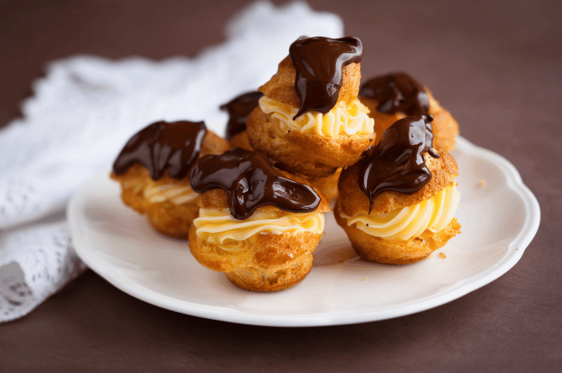 A small pile of profiteroles on a white scalloped plate, filled with swirls of piped yellow pastry cream and finished with a rich chocolate topping. A small pile of profiteroles on a white scalloped plate, filled with swirls of piped yellow pastry cream and finished with a rich chocolate topping.