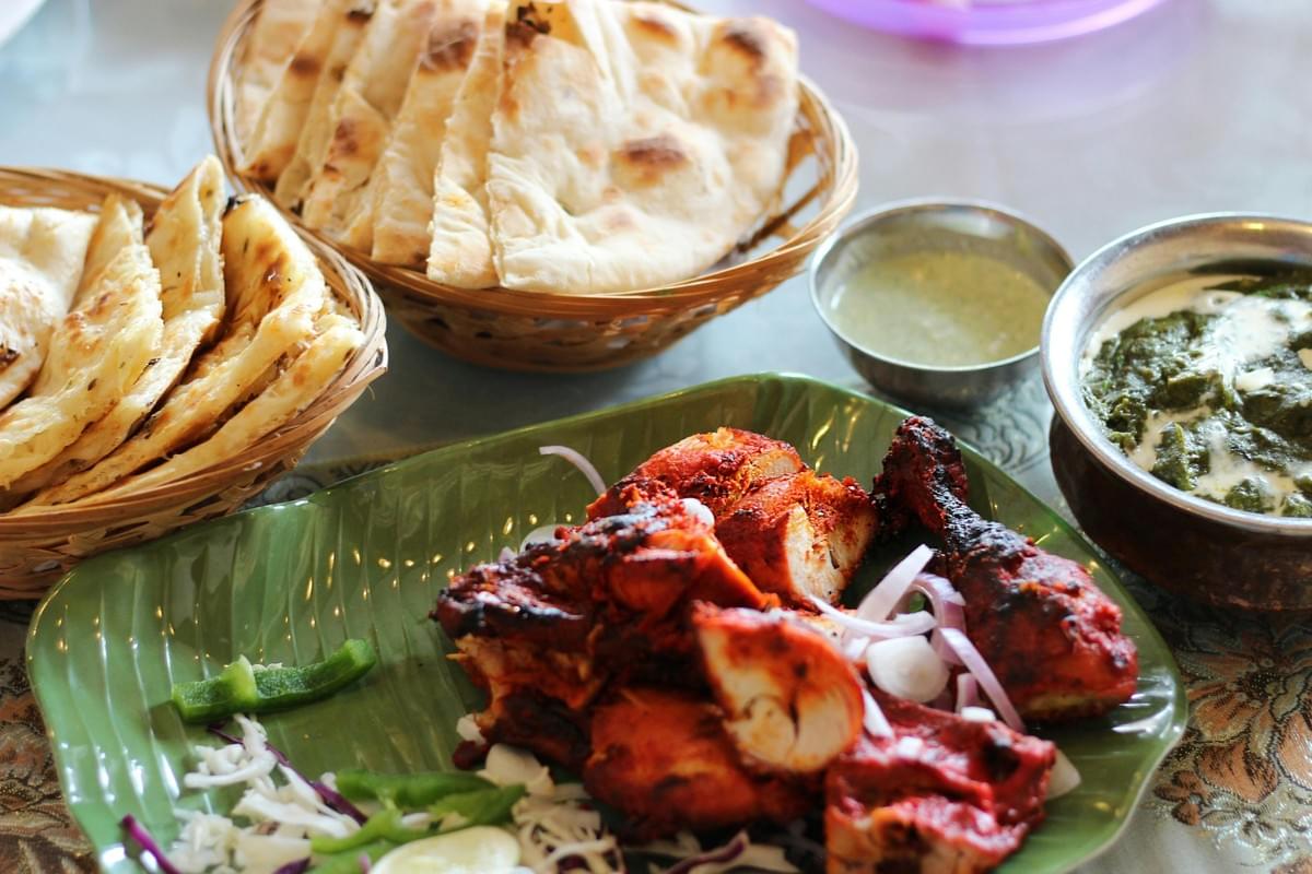A vibrant spread of Indian cuisine features bright red tandoori chicken served on a green leaf-patterned plate alongside a bowl of creamy palak paneer. Two woven baskets filled with warm naan bread and a small bowl of green chutney complete the table setting. A vibrant spread of Indian cuisine features bright red tandoori chicken served on a green leaf-patterned plate alongside a bowl of creamy palak paneer. Two woven baskets filled with warm naan bread and a small bowl of green chutney complete the table setting.