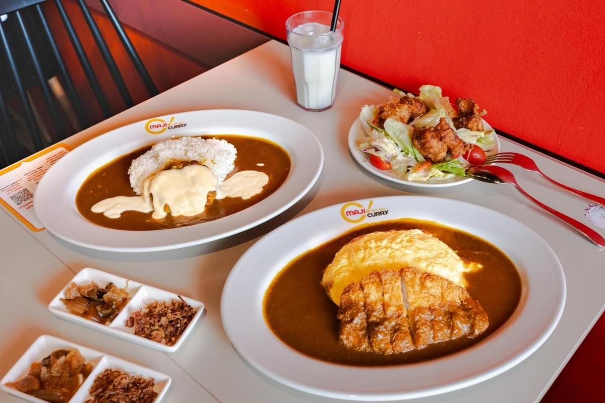 A high-angle shot features a table spread of Japanese curry dishes from Maji Curry, including an omurice curry with a crispy pork cutlet and another curry dish topped with melted cheese. Accompanying the main dishes are a side salad with fried chicken, a tall glass of a milky drink, and small side dishes of pickles and fried shallots. A high-angle shot features a table spread of Japanese curry dishes from Maji Curry, including an omurice curry with a crispy pork cutlet and another curry dish topped with melted cheese. Accompanying the main dishes are a side salad with fried chicken, a tall glass of a milky drink, and small side dishes of pickles and fried shallots.