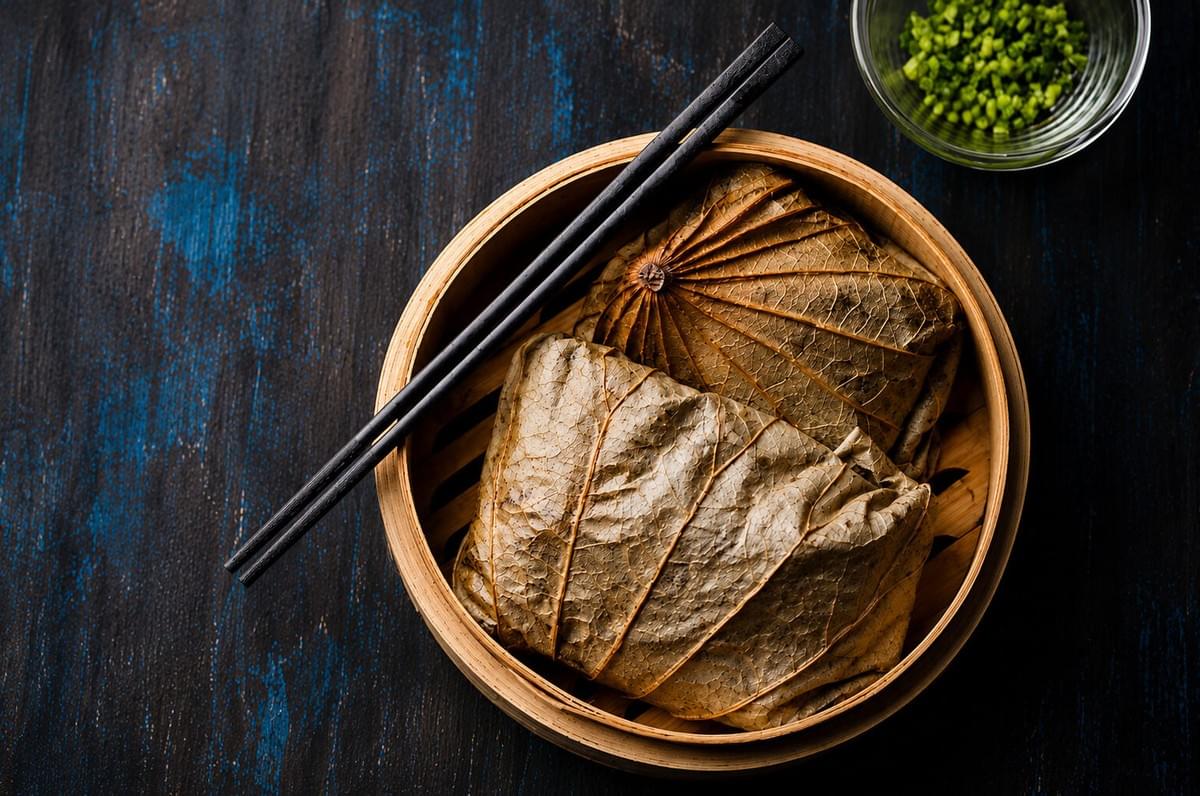 A top-down view of two dim sum parcels wrapped in dried lotus leaves inside a round bamboo steamer basket, with a pair of black chopsticks resting on top, set on a dark textured background. A top-down view of two dim sum parcels wrapped in dried lotus leaves inside a round bamboo steamer basket, with a pair of black chopsticks resting on top, set on a dark textured background.