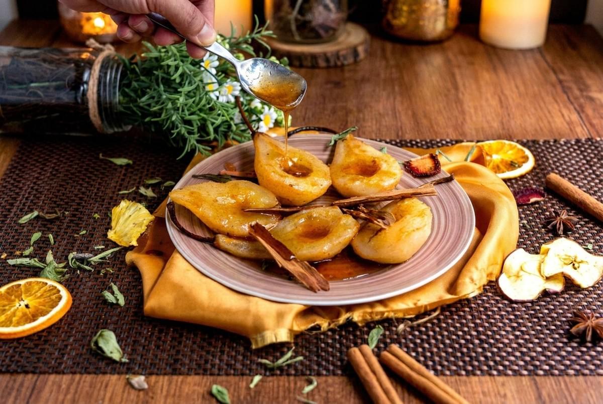 Halved poached pears arranged on a plate being drizzled with syrup from a spoon, garnished with cinnamon sticks. Halved poached pears arranged on a plate being drizzled with syrup from a spoon, garnished with cinnamon sticks.