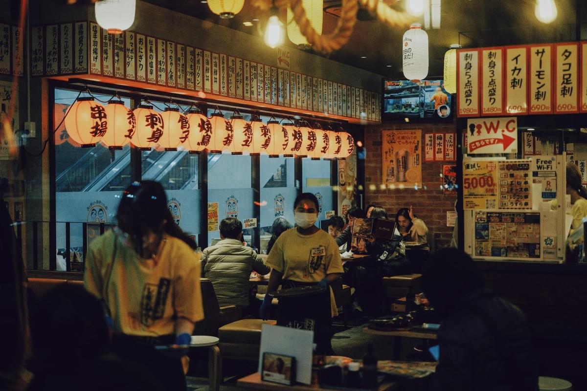 The interior of a bustling, warmly lit Japanese izakaya features traditional paper lanterns hanging overhead and walls covered in menu strips. Staff wearing face masks move between tables where customers are seated enjoying their meals. The interior of a bustling, warmly lit Japanese izakaya features traditional paper lanterns hanging overhead and walls covered in menu strips. Staff wearing face masks move between tables where customers are seated enjoying their meals.