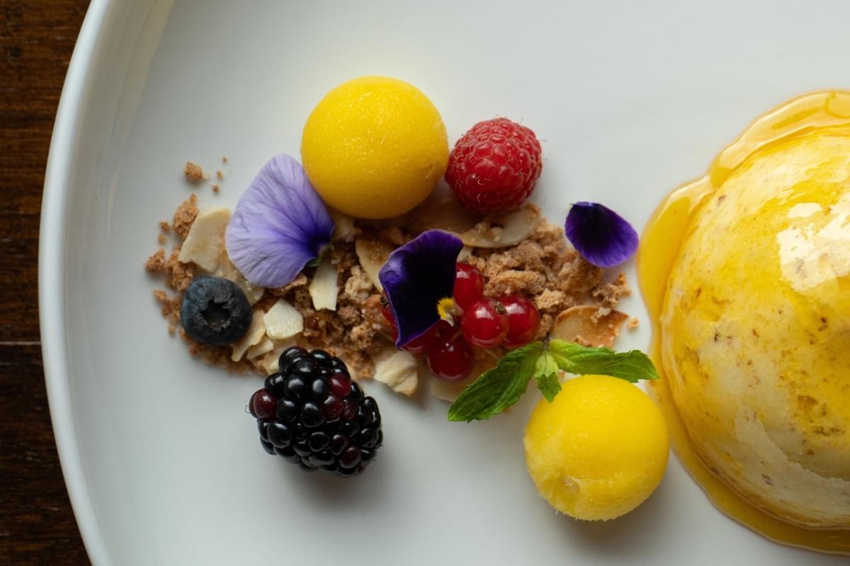 The image displays an elegantly plated gourmet dessert served on a white dish. It features a glazed round pastry draped in yellow syrup, paired alongside bright yellow sorbet scoops, fresh mixed berries, edible purple flower petals, and a bed of crisp crumble. The image displays an elegantly plated gourmet dessert served on a white dish. It features a glazed round pastry draped in yellow syrup, paired alongside bright yellow sorbet scoops, fresh mixed berries, edible purple flower petals, and a bed of crisp crumble.