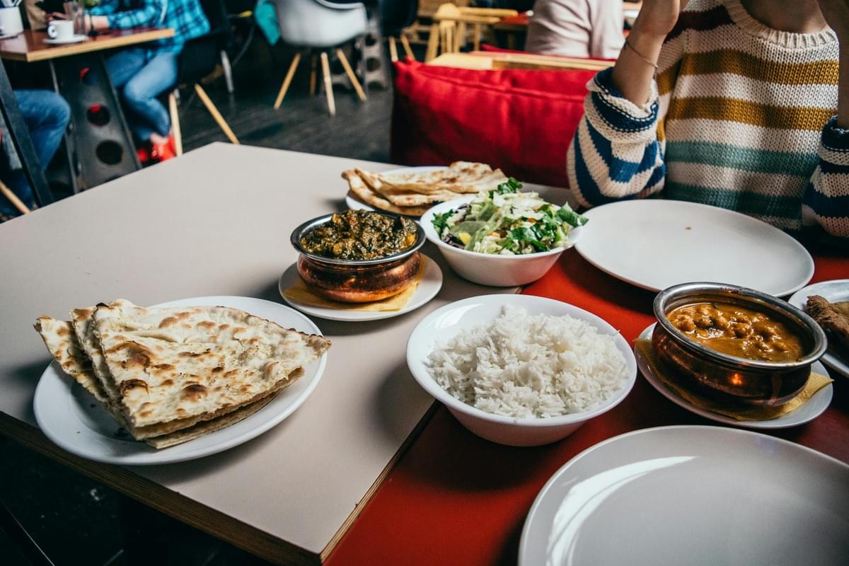 A table is spread with a variety of Indian dishes, including bowls of curry, white rice, a fresh green salad, and plates of naan bread. Two people are seated at the table in a restaurant setting, with empty white plates waiting to be served. A table is spread with a variety of Indian dishes, including bowls of curry, white rice, a fresh green salad, and plates of naan bread. Two people are seated at the table in a restaurant setting, with empty white plates waiting to be served.