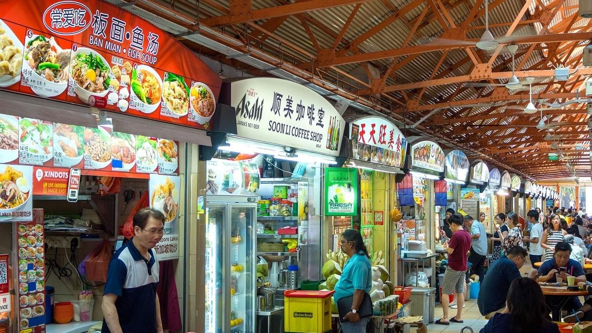 A vibrant indoor hawker centre in Southeast Asia features a row of colorful food stalls offering local dishes like Ban Mian and fish soup. Customers wander the narrow aisle or sit at nearby tables, creating a bustling atmosphere under the high, industrial-style ceiling. A vibrant indoor hawker centre in Southeast Asia features a row of colorful food stalls offering local dishes like Ban Mian and fish soup. Customers wander the narrow aisle or sit at nearby tables, creating a bustling atmosphere under the high, industrial-style ceiling.