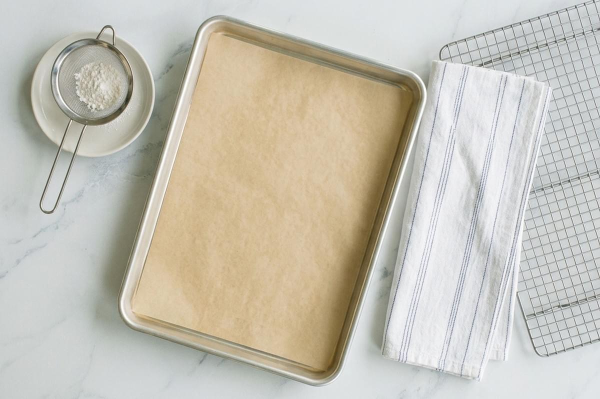 Baking sheet lined with parchment paper alongside a flour sifter, striped kitchen towel, and wire cooling rack ready for baking Baking sheet lined with parchment paper alongside a flour sifter, striped kitchen towel, and wire cooling rack ready for baking