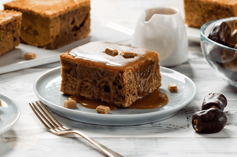A square slice of sticky toffee pudding generously coated in warm caramel sauce and topped with small fudge pieces, plated next to a bowl of fresh dates. A square slice of sticky toffee pudding generously coated in warm caramel sauce and topped with small fudge pieces, plated next to a bowl of fresh dates.