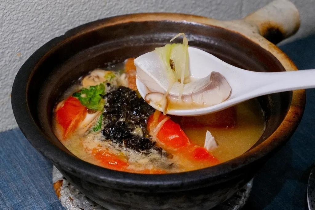 A white ceramic spoon scoops a piece of fish and shredded ginger from a simmering clay pot soup. The pot contains a rich broth filled with tomatoes, seaweed, and herbs. A white ceramic spoon scoops a piece of fish and shredded ginger from a simmering clay pot soup. The pot contains a rich broth filled with tomatoes, seaweed, and herbs.
