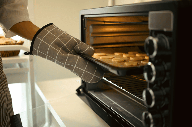 A baker wearing a grey checkered oven mitt carefully places a baking sheet of unbaked cookies into a fully preheated countertop oven, highlighting the importance of starting with the correct initial baking temperature to prevent spreading and ensure an even bake. A baker wearing a grey checkered oven mitt carefully places a baking sheet of unbaked cookies into a fully preheated countertop oven, highlighting the importance of starting with the correct initial baking temperature to prevent spreading and ensure an even bake.