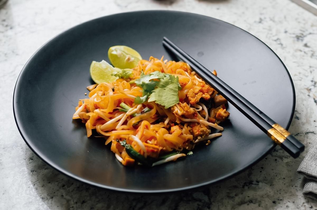 Close-up hero shot of authentic pad thai noodles plated on a black dish, garnished with bean sprouts, lime wedges, and fresh herbs, showcasing traditional Thai street food presentation. Close-up hero shot of authentic pad thai noodles plated on a black dish, garnished with bean sprouts, lime wedges, and fresh herbs, showcasing traditional Thai street food presentation.
