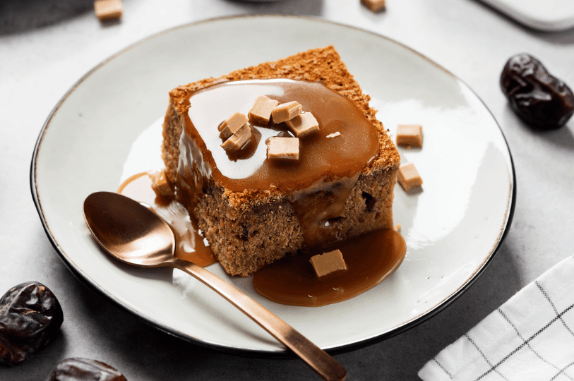 A close-up of a square cut of sticky toffee pudding drizzled with thick caramel and garnished with toffee squares, with a copper dessert spoon resting on the plate. A close-up of a square cut of sticky toffee pudding drizzled with thick caramel and garnished with toffee squares, with a copper dessert spoon resting on the plate.