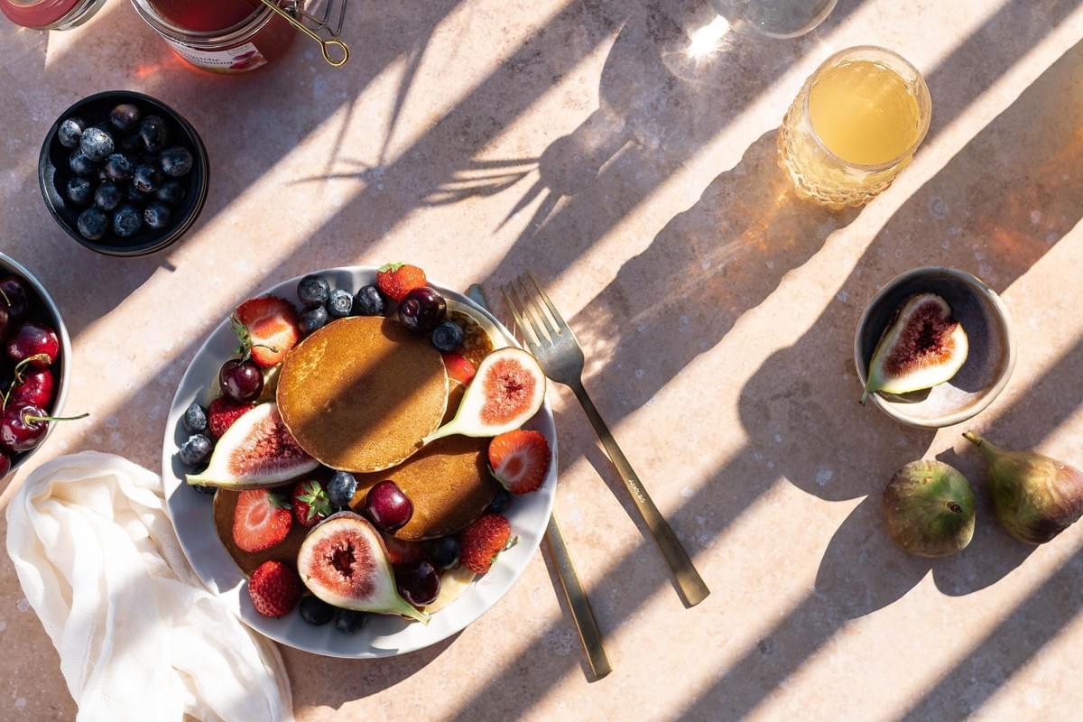 An overhead shot displays a plate of golden pancakes piled high with fresh strawberries, blueberries, cherries, and sliced figs on a sunlit stone table. Strong, linear shadows cast across the scene, which also includes gold utensils, a glass of juice, and bowls of extra fresh fruit. An overhead shot displays a plate of golden pancakes piled high with fresh strawberries, blueberries, cherries, and sliced figs on a sunlit stone table. Strong, linear shadows cast across the scene, which also includes gold utensils, a glass of juice, and bowls of extra fresh fruit.