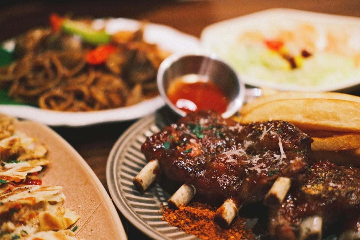A close-up shot features a plate of glazed barbecue ribs served with potato wedges and dipping sauce. Surrounding the main dish on the wooden table are additional plates containing pasta, quesadillas, and salad. A close-up shot features a plate of glazed barbecue ribs served with potato wedges and dipping sauce. Surrounding the main dish on the wooden table are additional plates containing pasta, quesadillas, and salad.
