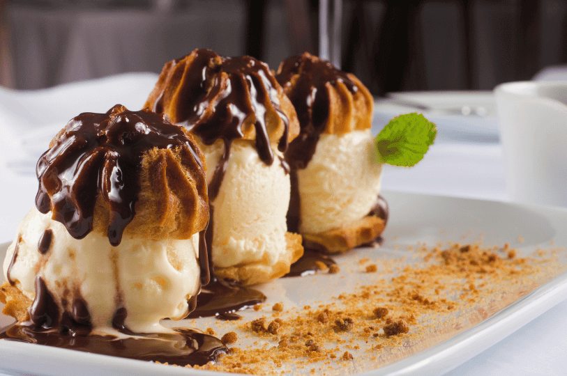 Three large profiteroles split open and stuffed with generous scoops of vanilla ice cream, heavily drizzled with chocolate sauce, and served on a white rectangular plate scattered with biscuit crumbs. Three large profiteroles split open and stuffed with generous scoops of vanilla ice cream, heavily drizzled with chocolate sauce, and served on a white rectangular plate scattered with biscuit crumbs.