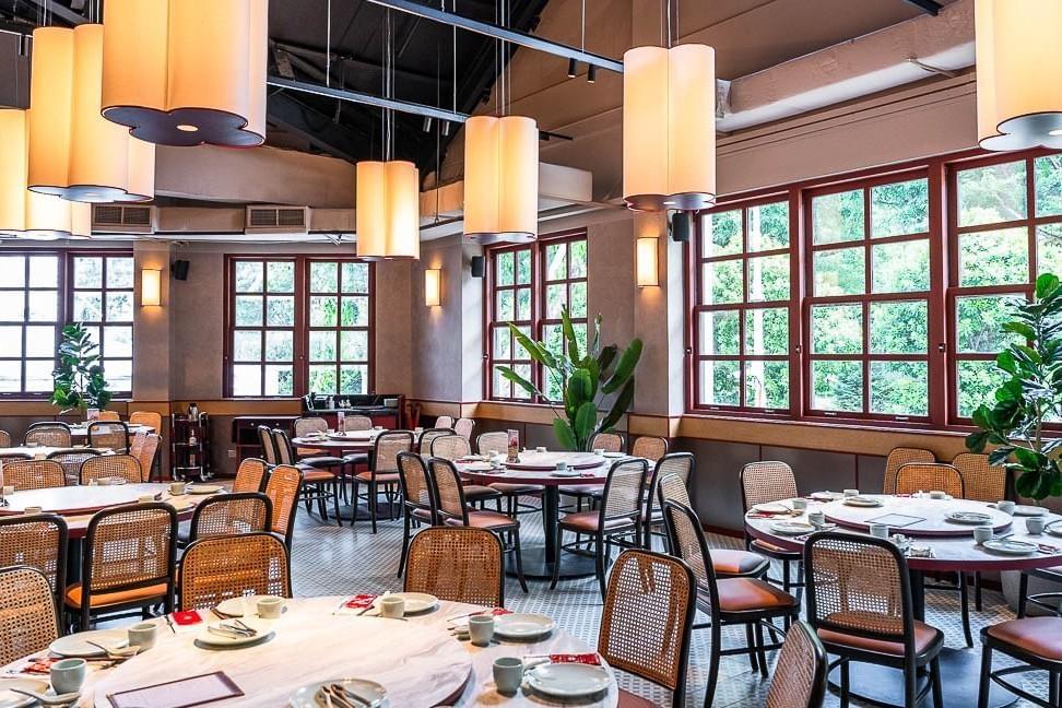 The interior of a spacious restaurant features round dining tables set with tableware, surrounded by woven-back chairs. Warm, glowing lantern-style pendant lights hang from the high ceiling, while large red-framed windows look out onto lush greenery. The interior of a spacious restaurant features round dining tables set with tableware, surrounded by woven-back chairs. Warm, glowing lantern-style pendant lights hang from the high ceiling, while large red-framed windows look out onto lush greenery.