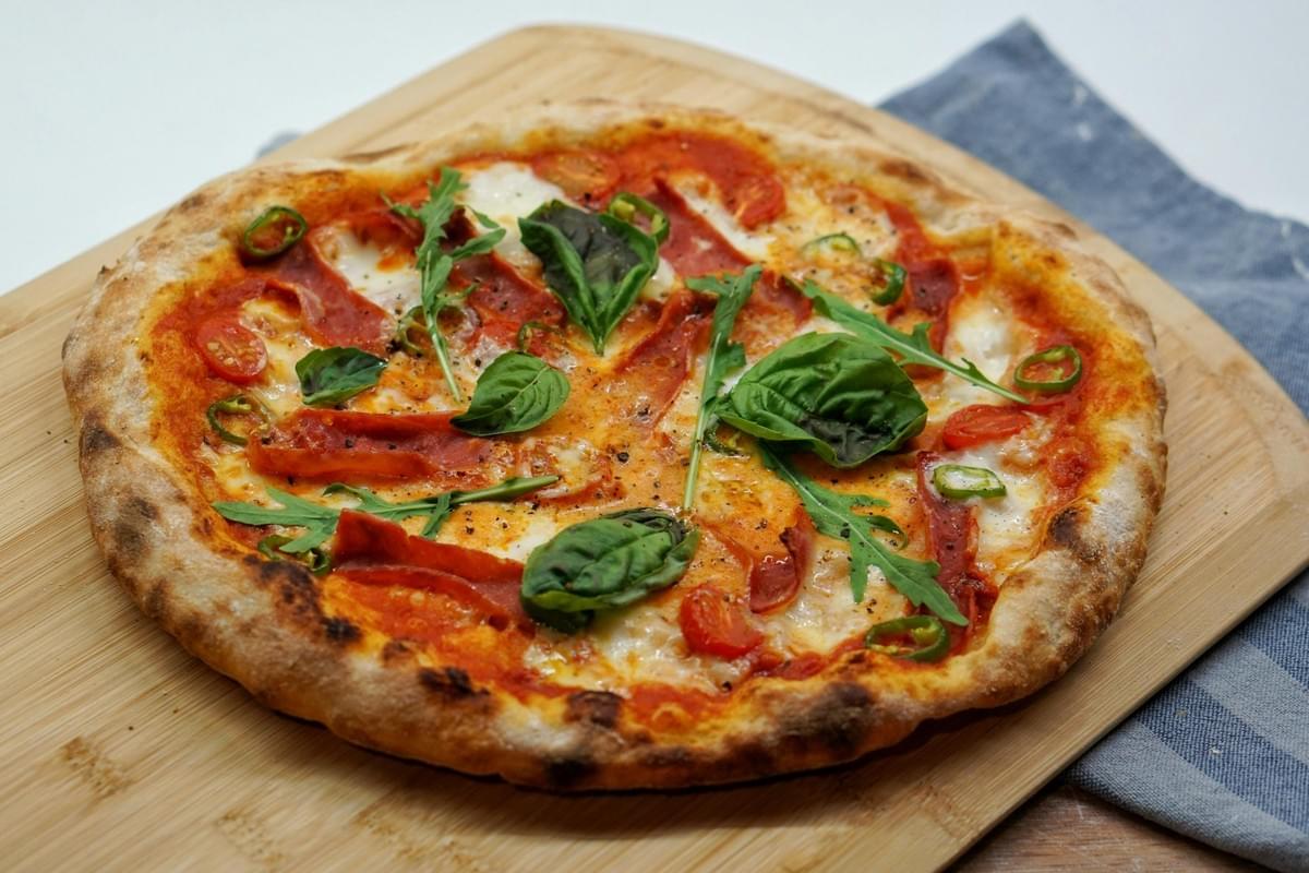 A freshly baked, rustic pizza with a charred crust sits on a wooden cutting board, topped with tomato sauce, melted mozzarella, and slices of cured meat. It is garnished with fresh basil leaves, arugula, cherry tomato halves, and sliced green chili peppers. A freshly baked, rustic pizza with a charred crust sits on a wooden cutting board, topped with tomato sauce, melted mozzarella, and slices of cured meat. It is garnished with fresh basil leaves, arugula, cherry tomato halves, and sliced green chili peppers.