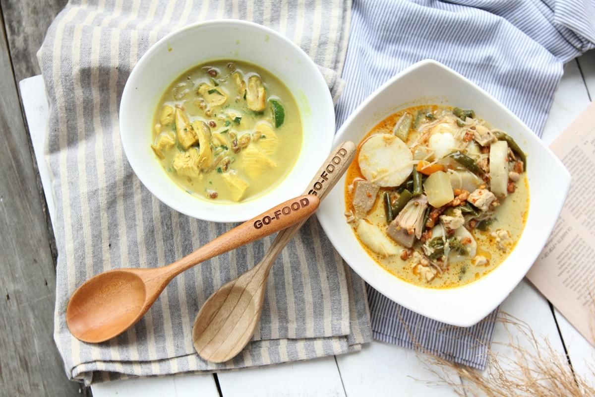 The image displays two white bowls of flavorful Indonesian curry dishes resting on striped napkins and a wooden surface. Two wooden spoons, one branded with "GO-FOOD," are placed between the bowls of what appears to be Opor Ayam and Lontong Sayur. The image displays two white bowls of flavorful Indonesian curry dishes resting on striped napkins and a wooden surface. Two wooden spoons, one branded with "GO-FOOD," are placed between the bowls of what appears to be Opor Ayam and Lontong Sayur.