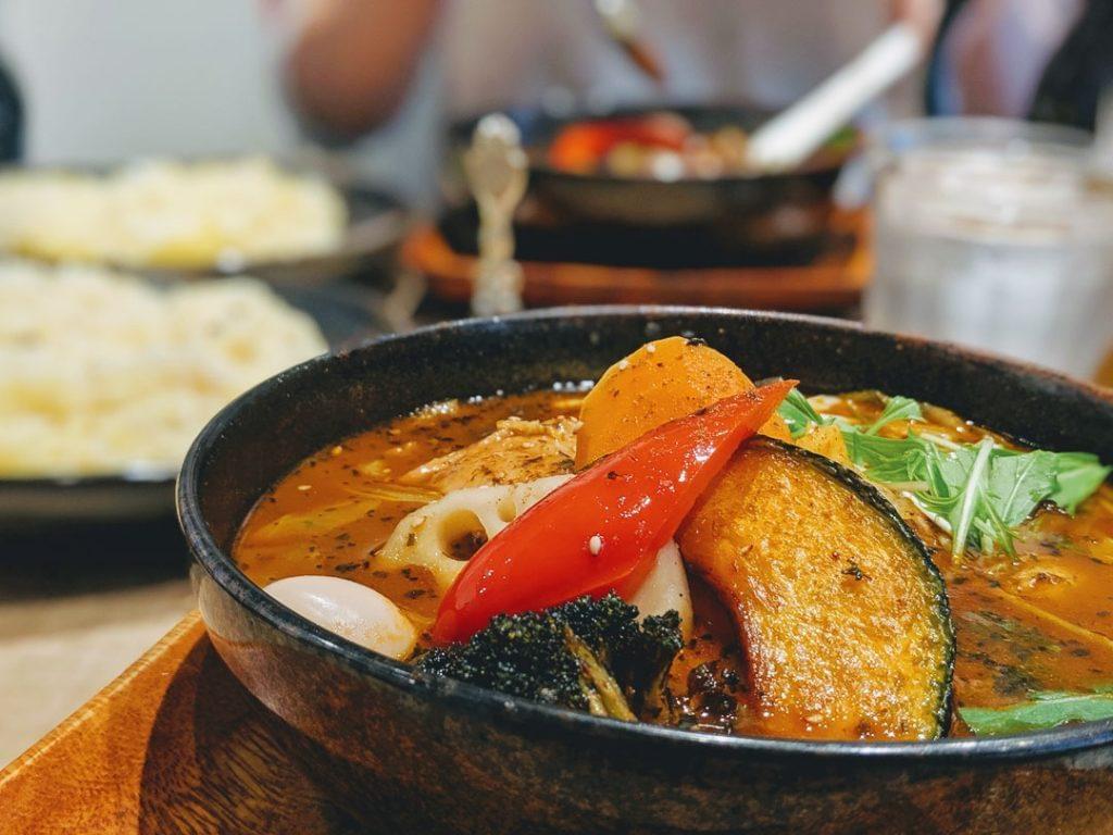 A close-up shot features a bowl of Japanese soup curry filled with rich, spiced broth and vibrant toppings like roasted pumpkin, red bell pepper, broccoli, and lotus root. In the blurred background, another diner and side dishes of rice complete the cozy restaurant setting. A close-up shot features a bowl of Japanese soup curry filled with rich, spiced broth and vibrant toppings like roasted pumpkin, red bell pepper, broccoli, and lotus root. In the blurred background, another diner and side dishes of rice complete the cozy restaurant setting.