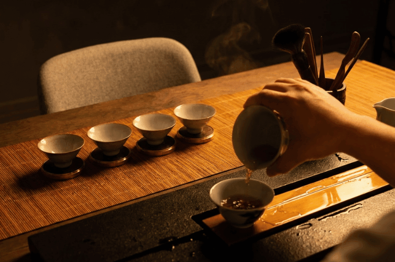 The tea master's hand gently pours golden tea from a white ceramic vessel into a line of small tasting cups, with soft steam rising in the warm background at the Tea Room by Ki-setsu. The tea master's hand gently pours golden tea from a white ceramic vessel into a line of small tasting cups, with soft steam rising in the warm background at the Tea Room by Ki-setsu.