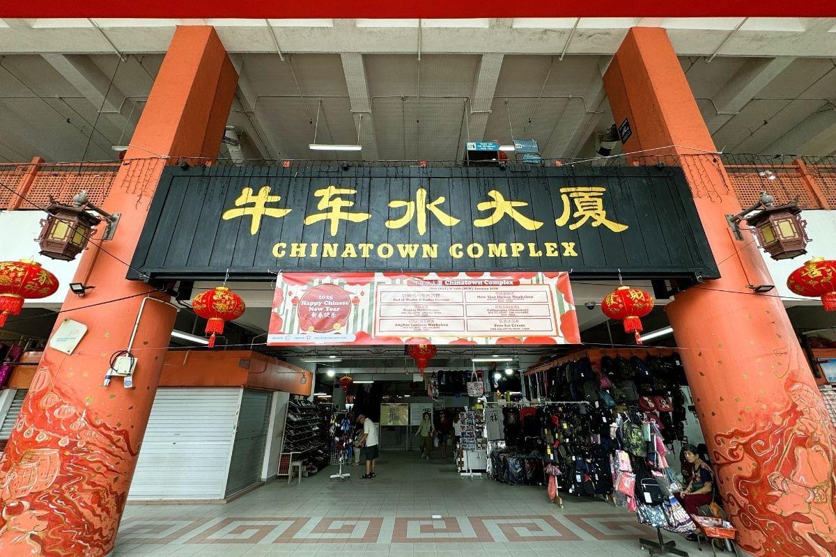 The entrance to Chinatown Complex features a prominent black sign with gold lettering and festive red lanterns hanging from large orange pillars. A colorful banner celebrating the 2025 Happy Chinese New Year is displayed below the sign, overlooking stalls filled with bags and merchandise. The entrance to Chinatown Complex features a prominent black sign with gold lettering and festive red lanterns hanging from large orange pillars. A colorful banner celebrating the 2025 Happy Chinese New Year is displayed below the sign, overlooking stalls filled with bags and merchandise.