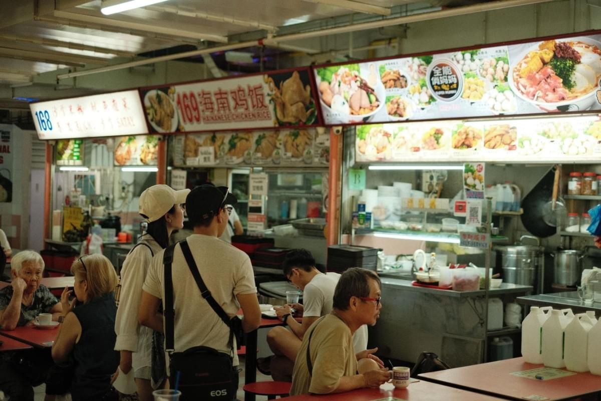 A bustling hawker center filled with colorful food stall signs displaying menus in Chinese and English. People are seated at red dining tables, while others stand and wait for their orders under bright overhead lights. A bustling hawker center filled with colorful food stall signs displaying menus in Chinese and English. People are seated at red dining tables, while others stand and wait for their orders under bright overhead lights.