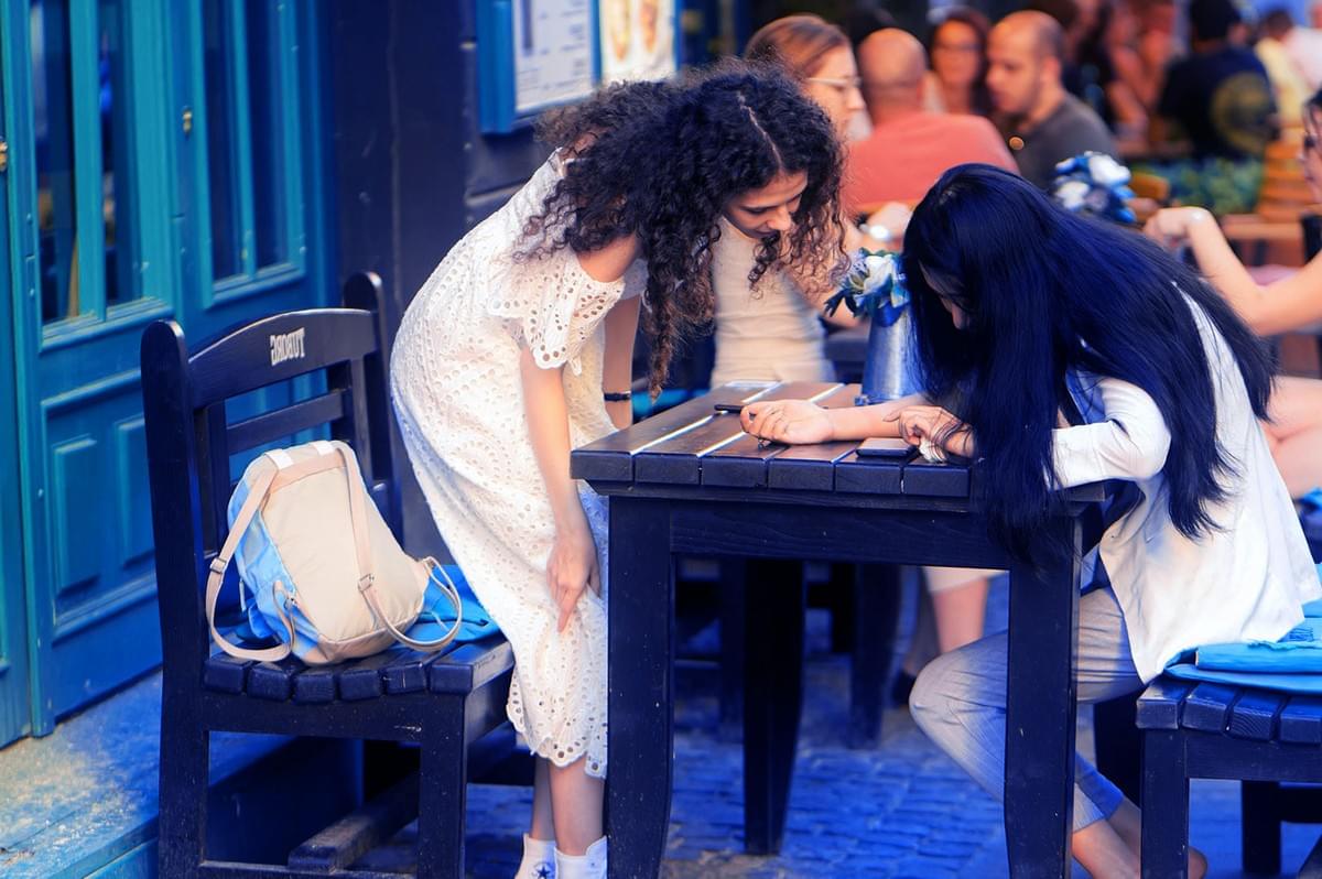 Two women are engaged in conversation at an outdoor café. One stands while the other sits, both focused on something on a smartphone. Relaxed atmosphere. Two women are engaged in conversation at an outdoor café. One stands while the other sits, both focused on something on a smartphone. Relaxed atmosphere.