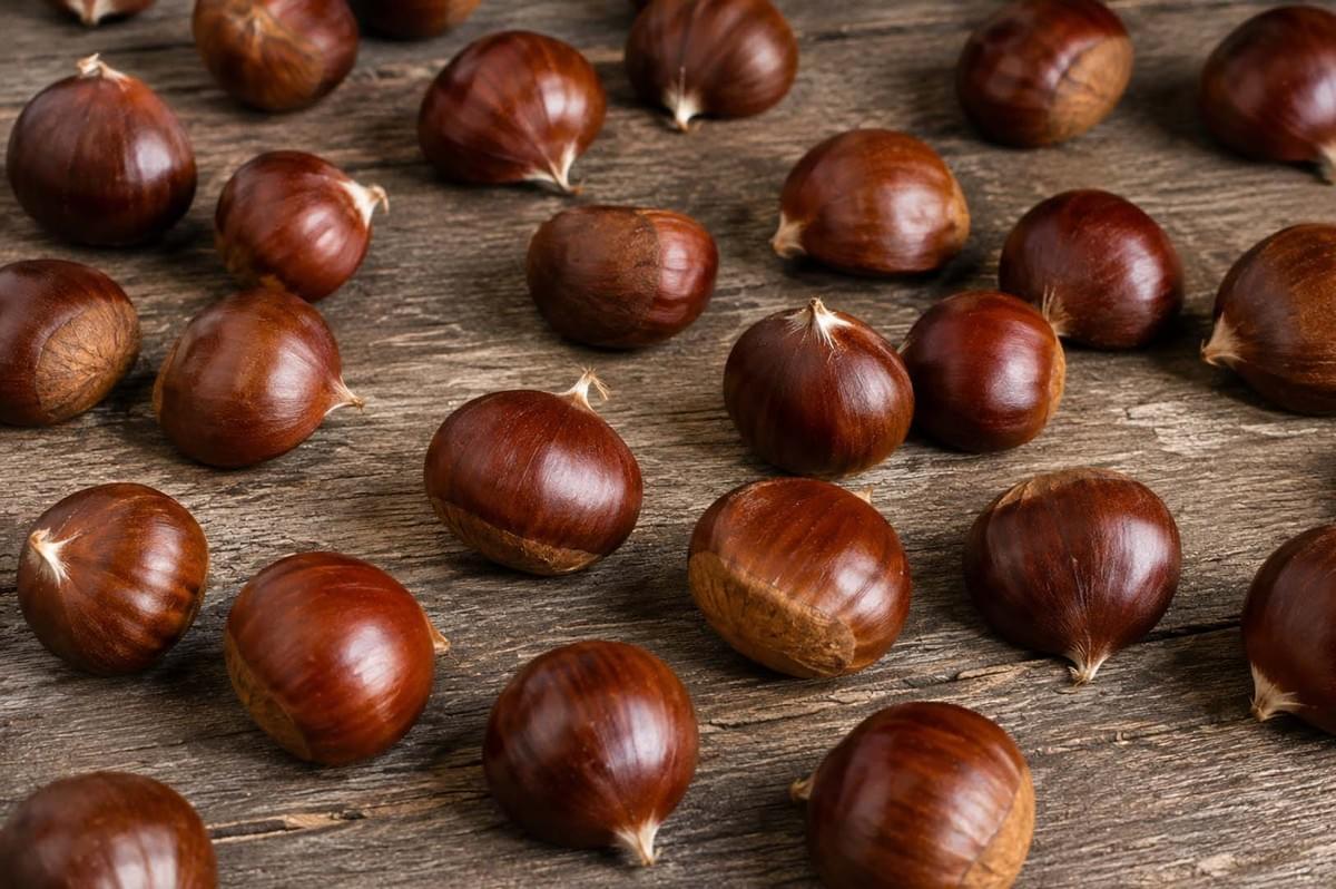 A spread of raw, shiny brown chestnuts scattered across a rustic wooden table surface A spread of raw, shiny brown chestnuts scattered across a rustic wooden table surface