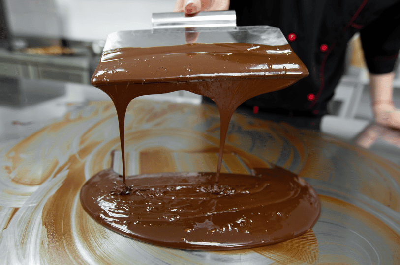 Close-up of a chocolatier using a wide metal bench scraper to lift and pour glossy melted chocolate, creating thick ribbons as it falls back onto a smooth work surface during the table tempering process. Close-up of a chocolatier using a wide metal bench scraper to lift and pour glossy melted chocolate, creating thick ribbons as it falls back onto a smooth work surface during the table tempering process.