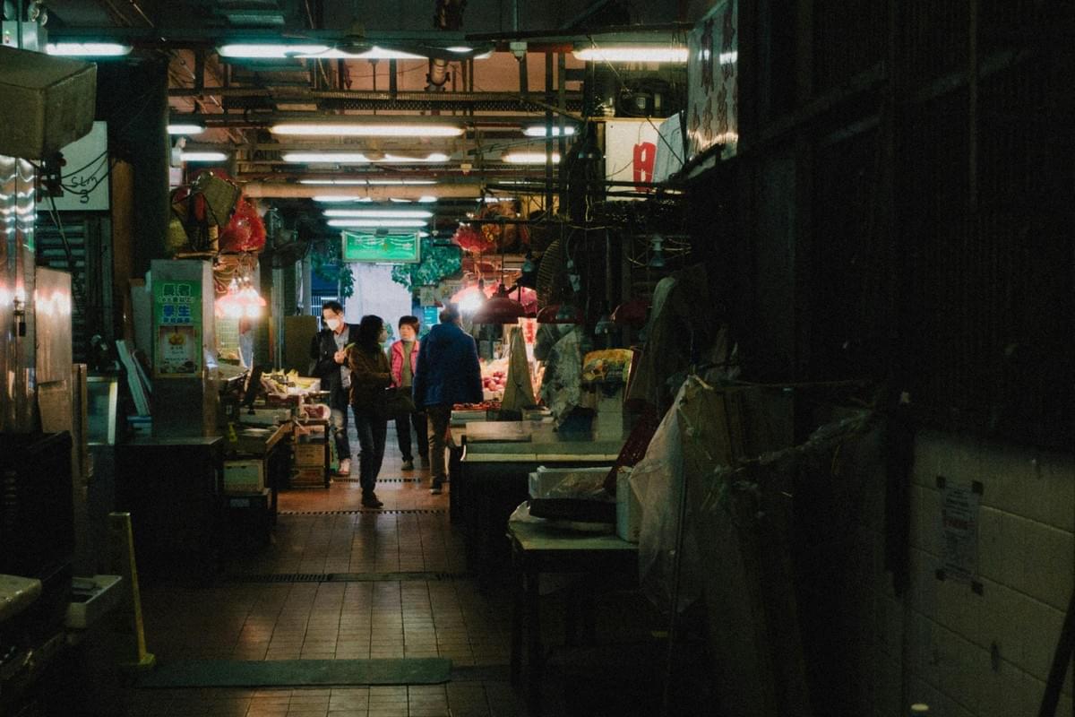 Under the cool glow of overhead fluorescent lights, a small group of people walks away down a dark, tiled corridor within an indoor market. The aisle is lined with stalls featuring stacked boxes and hanging red fixtures, creating a gritty, atmospheric scene with exposed pipes running along the ceiling. Under the cool glow of overhead fluorescent lights, a small group of people walks away down a dark, tiled corridor within an indoor market. The aisle is lined with stalls featuring stacked boxes and hanging red fixtures, creating a gritty, atmospheric scene with exposed pipes running along the ceiling.