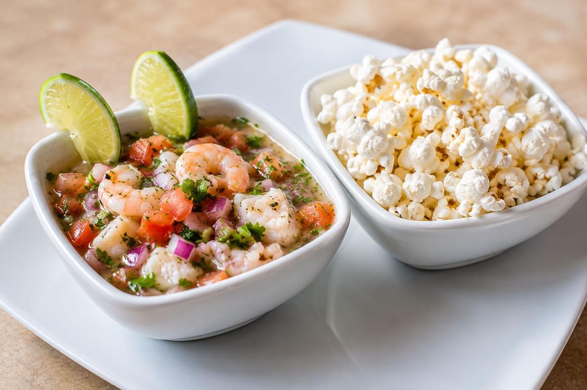 White bowl of shrimp ceviche garnished with lime wedges served alongside a bowl of popcorn on a white plate White bowl of shrimp ceviche garnished with lime wedges served alongside a bowl of popcorn on a white plate