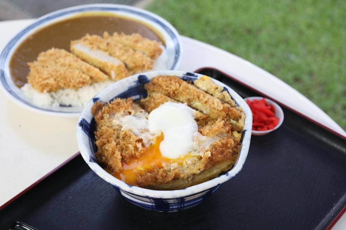 A blue and white bowl of katsudon sits on a black tray, featuring crispy golden-brown tonkatsu topped with a soft-boiled egg with a runny yolk. In the background, a second plate presents katsu curry with sliced fried pork cutlets over white rice and thick Japanese curry sauce. A blue and white bowl of katsudon sits on a black tray, featuring crispy golden-brown tonkatsu topped with a soft-boiled egg with a runny yolk. In the background, a second plate presents katsu curry with sliced fried pork cutlets over white rice and thick Japanese curry sauce.