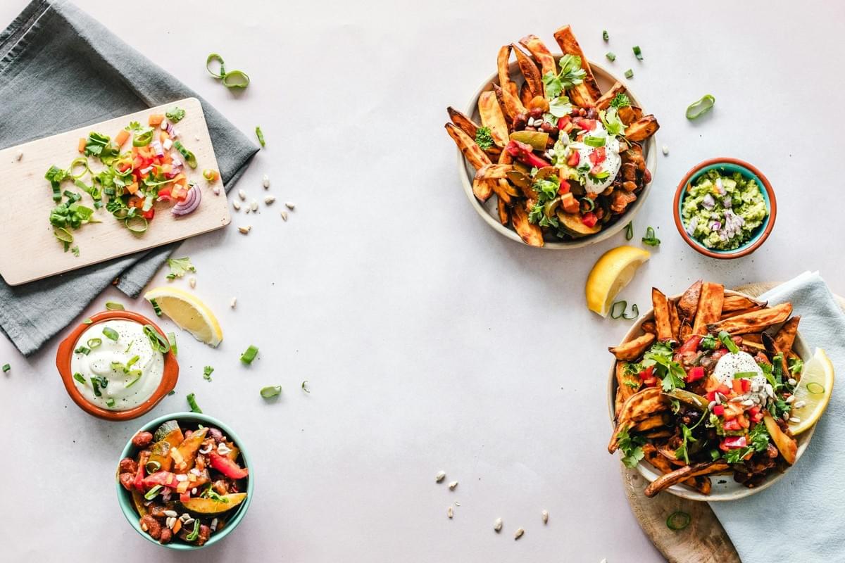 The image shows a vibrant top-down view of sweet potato wedges topped with chili, fresh herbs, and various garnishes. Surrounding the main plates are small bowls containing salsa, guacamole, and sour cream, alongside a cutting board with freshly chopped vegetables. The image shows a vibrant top-down view of sweet potato wedges topped with chili, fresh herbs, and various garnishes. Surrounding the main plates are small bowls containing salsa, guacamole, and sour cream, alongside a cutting board with freshly chopped vegetables.