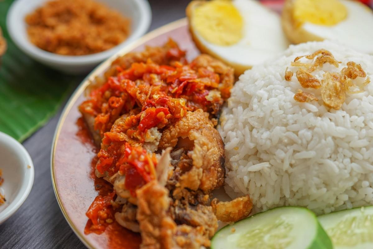 A plate of white rice topped with fried shallots is served alongside crispy fried chicken smothered in a vibrant, chunky red chili sambal. The meal is accompanied by sliced hard-boiled eggs and fresh cucumber rounds, with side bowls visible in the background. A plate of white rice topped with fried shallots is served alongside crispy fried chicken smothered in a vibrant, chunky red chili sambal. The meal is accompanied by sliced hard-boiled eggs and fresh cucumber rounds, with side bowls visible in the background.