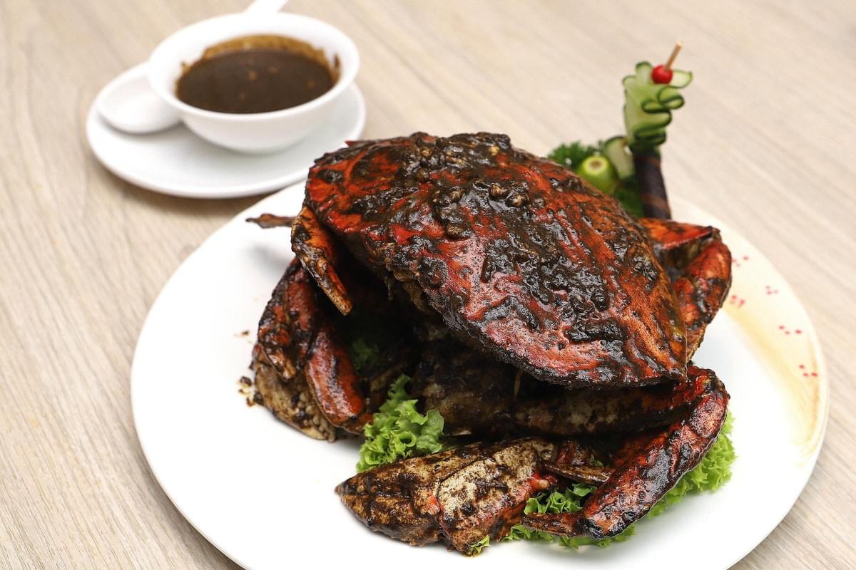 A whole cooked crab coated in dark black pepper sauce rests on a bed of lettuce on a large white plate. A small white bowl filled with extra sauce sits in the background on a wooden table. A whole cooked crab coated in dark black pepper sauce rests on a bed of lettuce on a large white plate. A small white bowl filled with extra sauce sits in the background on a wooden table.