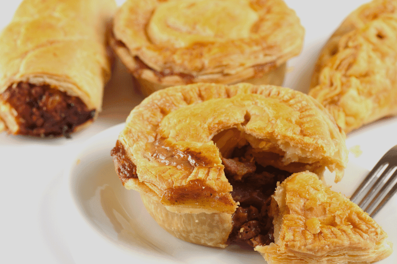 An assortment of Australian bakery favorites including a classic meat pie with a bite taken out, a sausage roll, and a pastie on a white plate. An assortment of Australian bakery favorites including a classic meat pie with a bite taken out, a sausage roll, and a pastie on a white plate.