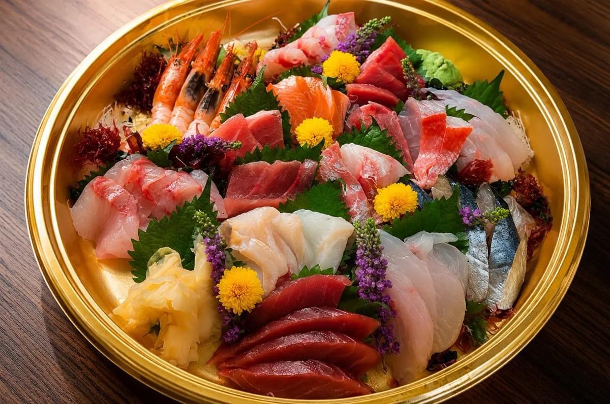 Close-up of a large gold platter generously packed with assorted raw fish slices, prawns, wasabi, and floral garnishes. Close-up of a large gold platter generously packed with assorted raw fish slices, prawns, wasabi, and floral garnishes.