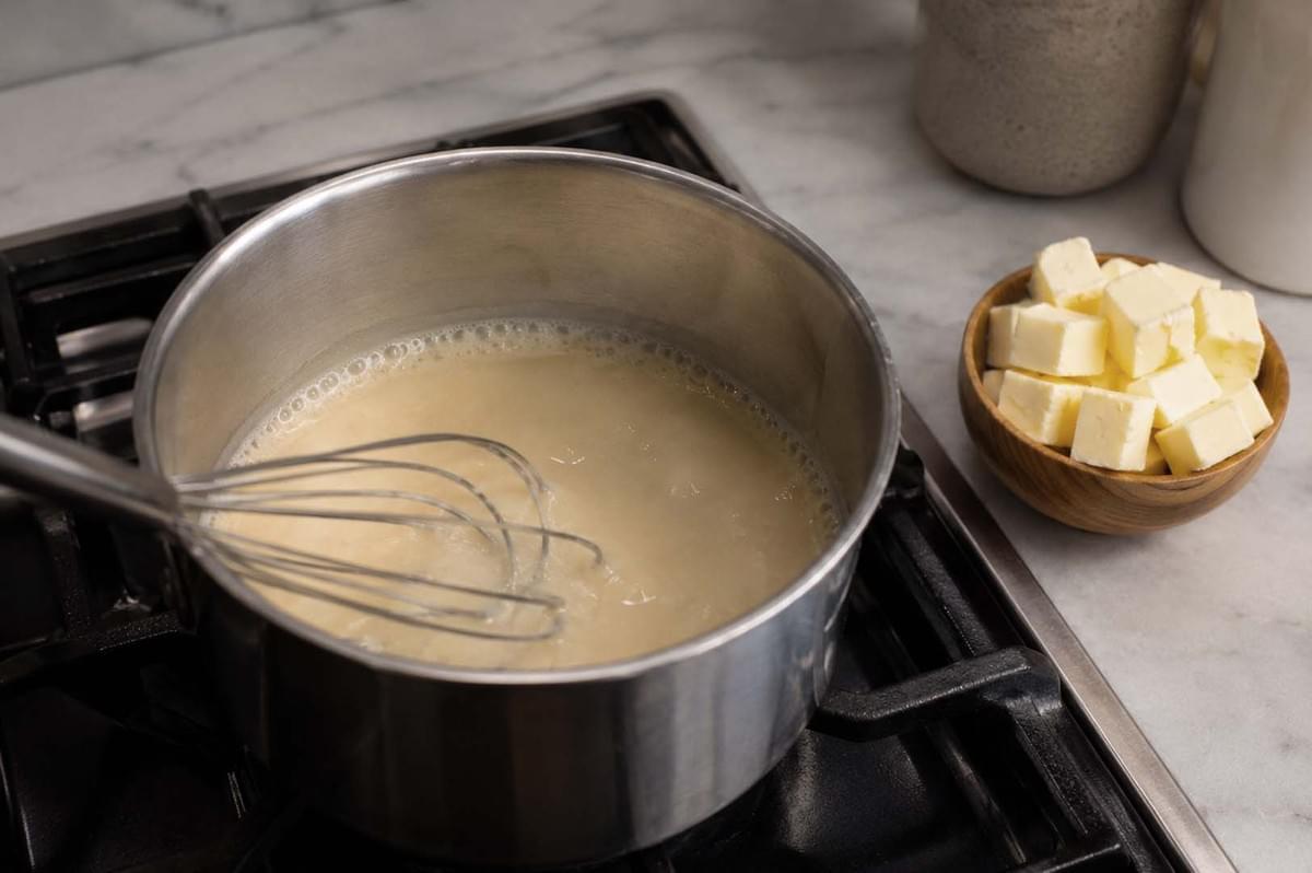 Whisk stirring a creamy sauce in a stainless steel pot on a stovetop, with cubed butter nearby Whisk stirring a creamy sauce in a stainless steel pot on a stovetop, with cubed butter nearby