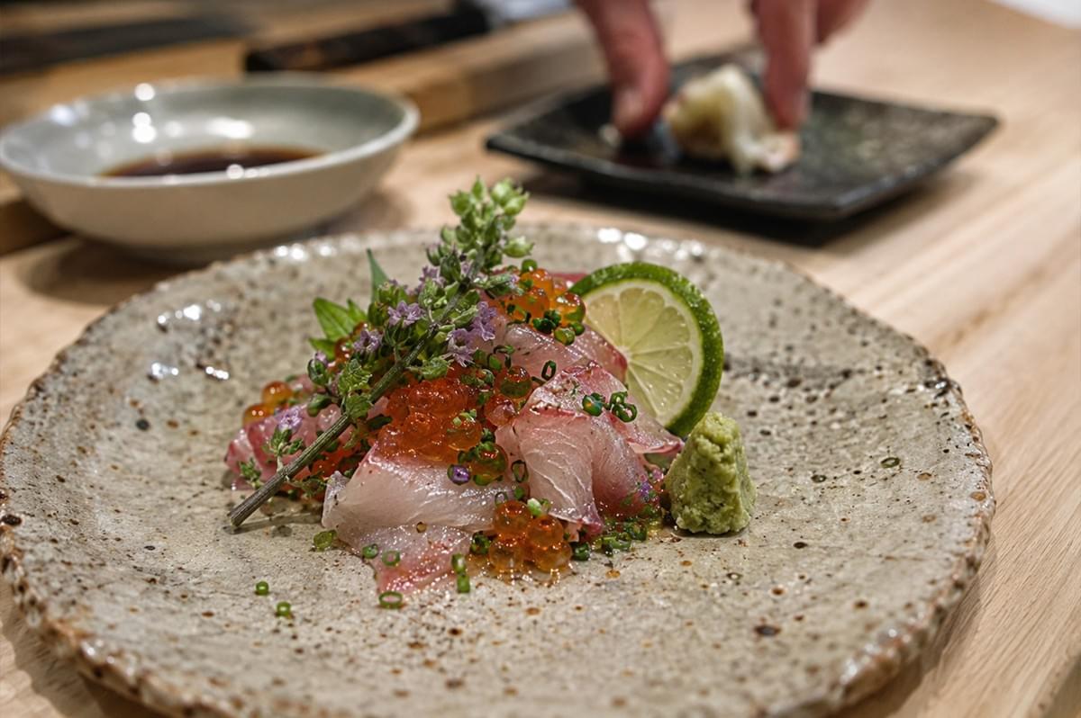A plate of sliced raw fish, or sashimi, is artfully arranged on a textured, speckled ceramic plate. The fish is topped with bright orange salmon roe, finely chopped green chives, and a sprig of purple-flowering basil. Beside the fish sits a neat dollop of bright green wasabi, and a slice of lime adds a vibrant accent to the composition, all set against a light-colored wooden workspace. A plate of sliced raw fish, or sashimi, is artfully arranged on a textured, speckled ceramic plate. The fish is topped with bright orange salmon roe, finely chopped green chives, and a sprig of purple-flowering basil. Beside the fish sits a neat dollop of bright green wasabi, and a slice of lime adds a vibrant accent to the composition, all set against a light-colored wooden workspace.