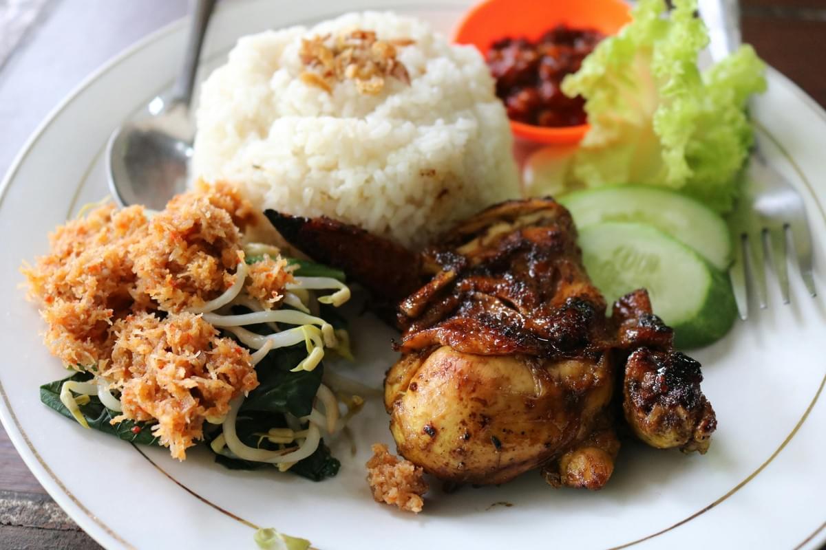 A plate of Indonesian food features a mound of white rice topped with fried shallots alongside a piece of grilled chicken and a serving of *urap* salad made of bean sprouts, greens, and spiced grated coconut. The dish is garnished with sliced cucumbers, lettuce, and a small orange bowl of red *sambal* chili paste. A plate of Indonesian food features a mound of white rice topped with fried shallots alongside a piece of grilled chicken and a serving of *urap* salad made of bean sprouts, greens, and spiced grated coconut. The dish is garnished with sliced cucumbers, lettuce, and a small orange bowl of red *sambal* chili paste.