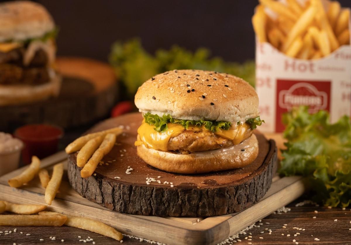 A close-up shot features a chicken burger topped with melted cheese and lettuce on a rustic wooden serving board, sprinkled with sesame seeds. In the background, a box of golden french fries and another burger are softly blurred on a dark, moody tabletop. A close-up shot features a chicken burger topped with melted cheese and lettuce on a rustic wooden serving board, sprinkled with sesame seeds. In the background, a box of golden french fries and another burger are softly blurred on a dark, moody tabletop.