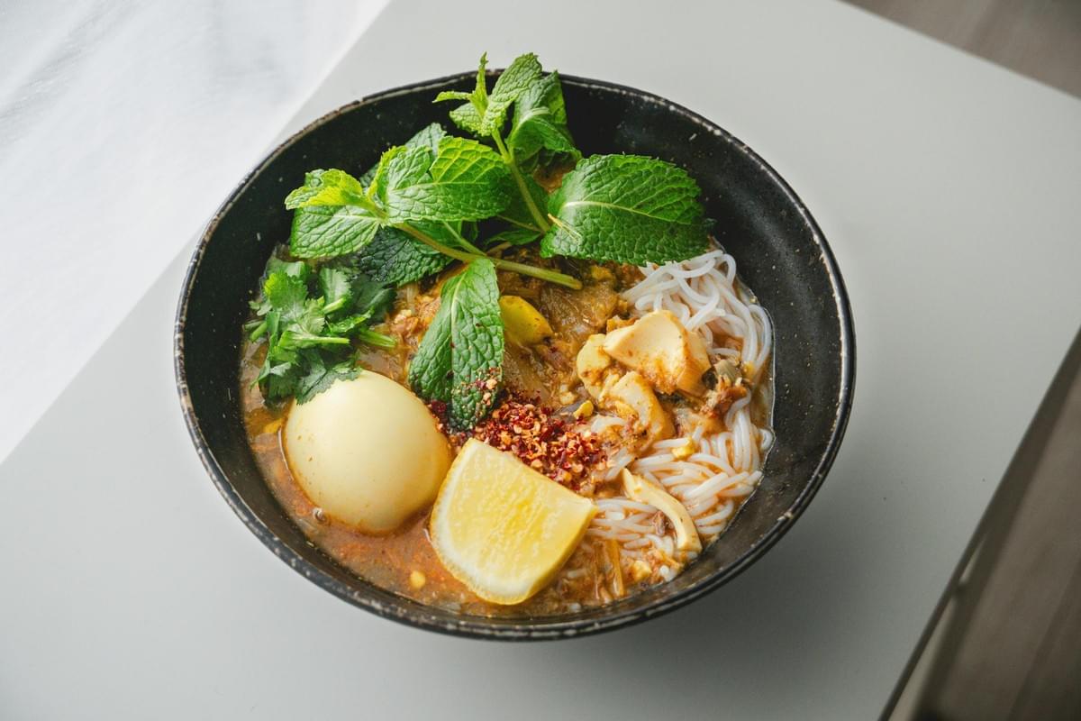 A bowl of Mohinga, a Burmese rice noodle and fish soup, is garnished with a hard-boiled egg, fresh mint, coriander, and a wedge of lemon. The dish is presented in a dark, textured ceramic bowl resting on a clean, white surface. A bowl of Mohinga, a Burmese rice noodle and fish soup, is garnished with a hard-boiled egg, fresh mint, coriander, and a wedge of lemon. The dish is presented in a dark, textured ceramic bowl resting on a clean, white surface.