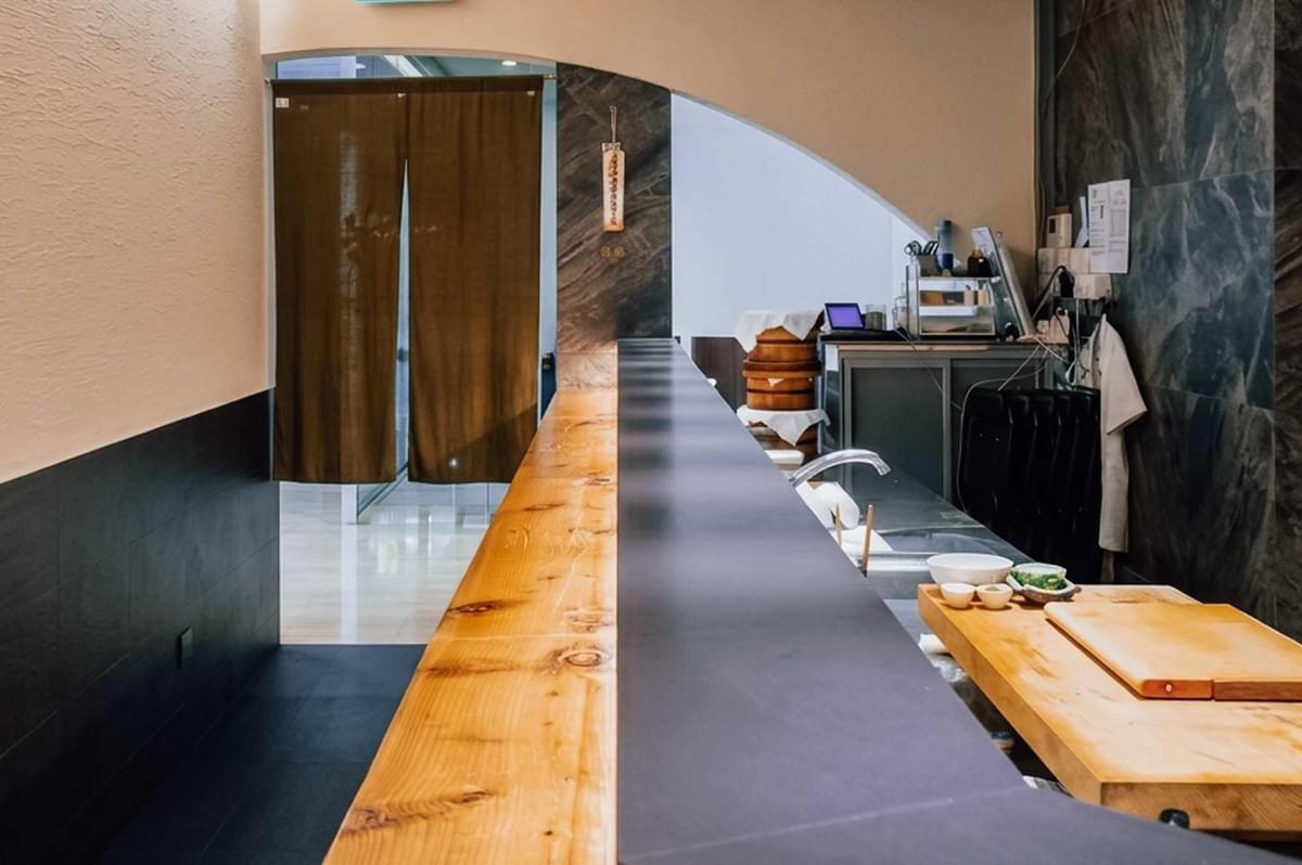A perspective view along the sleek wooden counter of a standing sushi bar, showing the clean preparation station equipped with cutting boards on the right and a brown entryway curtain hanging at the far end of the hallway. A perspective view along the sleek wooden counter of a standing sushi bar, showing the clean preparation station equipped with cutting boards on the right and a brown entryway curtain hanging at the far end of the hallway.