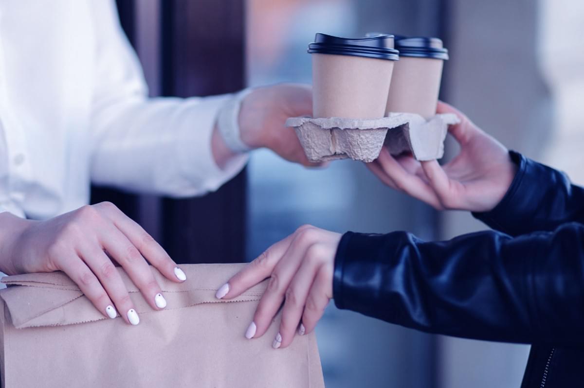 A person in a white shirt hands over a paper bag and a tray of coffee cups to another in a black jacket, suggesting a coffee takeout exchange. A person in a white shirt hands over a paper bag and a tray of coffee cups to another in a black jacket, suggesting a coffee takeout exchange.