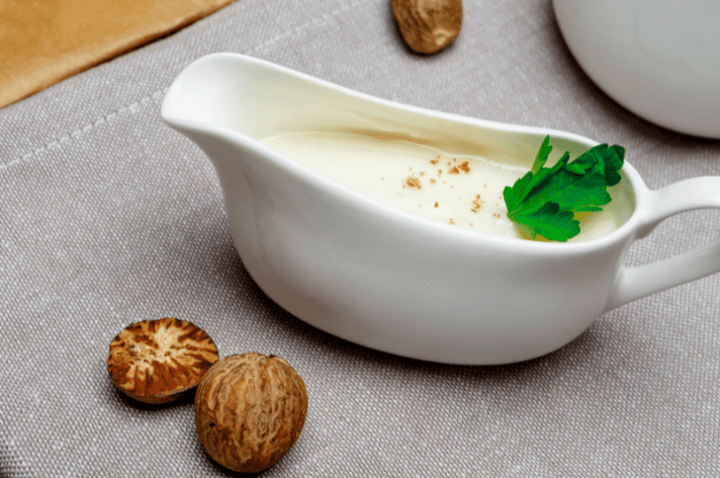 A close-up of a white ceramic gravy boat filled with smooth, creamy Béchamel sauce, garnished with a pinch of nutmeg and a fresh parsley sprig, with whole nutmeg seeds resting on a grey linen napkin nearby. A close-up of a white ceramic gravy boat filled with smooth, creamy Béchamel sauce, garnished with a pinch of nutmeg and a fresh parsley sprig, with whole nutmeg seeds resting on a grey linen napkin nearby.