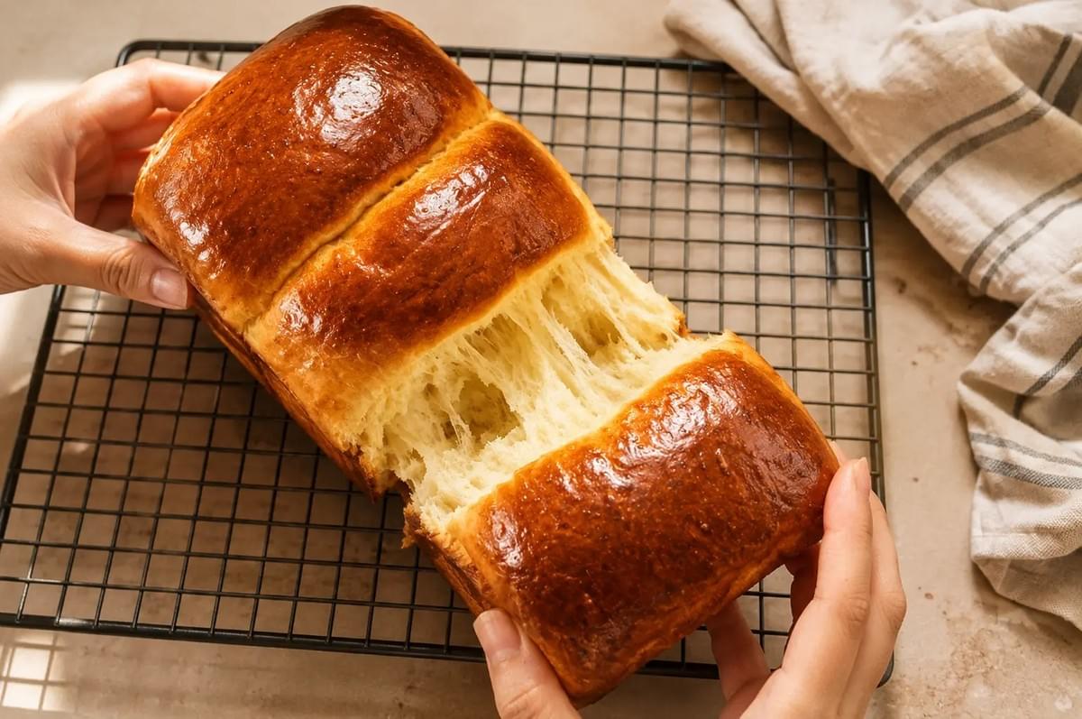 Hands gently pulling apart a soft, freshly baked brioche loaf over a wire cooling rack. Hands gently pulling apart a soft, freshly baked brioche loaf over a wire cooling rack.