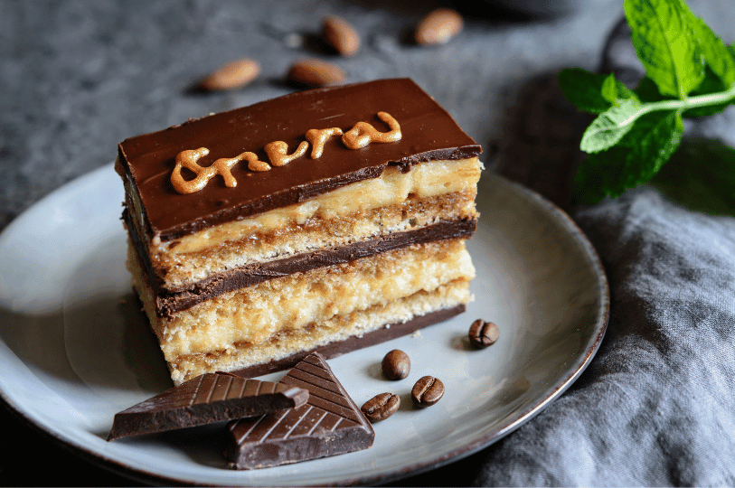 A decadent slice of layered opera cake on a grey plate featuring the word "Opera" beautifully piped in gold across the top chocolate glaze, surrounded by chocolate shards, coffee beans, and fresh mint. A decadent slice of layered opera cake on a grey plate featuring the word "Opera" beautifully piped in gold across the top chocolate glaze, surrounded by chocolate shards, coffee beans, and fresh mint.